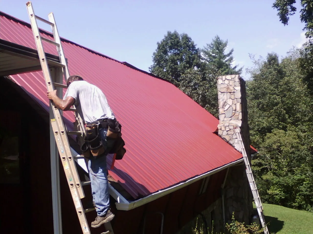 Roofer on a ladder beside a red metal roof and stone chimney — Soffit & Fascia Repair by Roofing Brothers in Galway